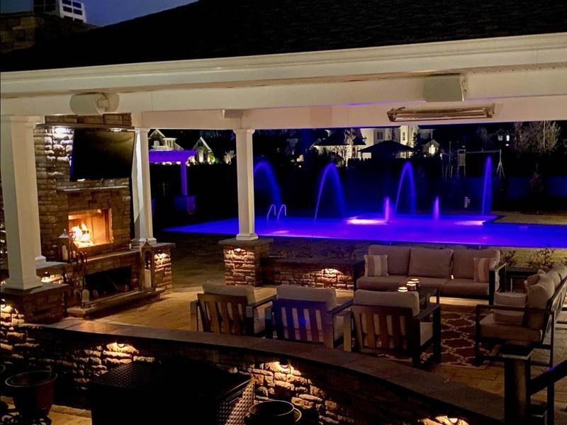 Outdoor patio with fireplace, seating, and illuminated pool fountains at night.