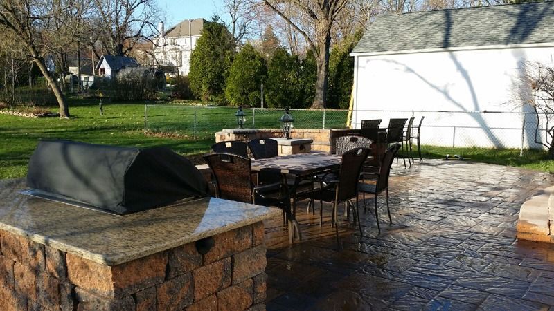 Outdoor patio with built-in grill, dining table, chairs, and surrounding lawn. Brick and granite features.