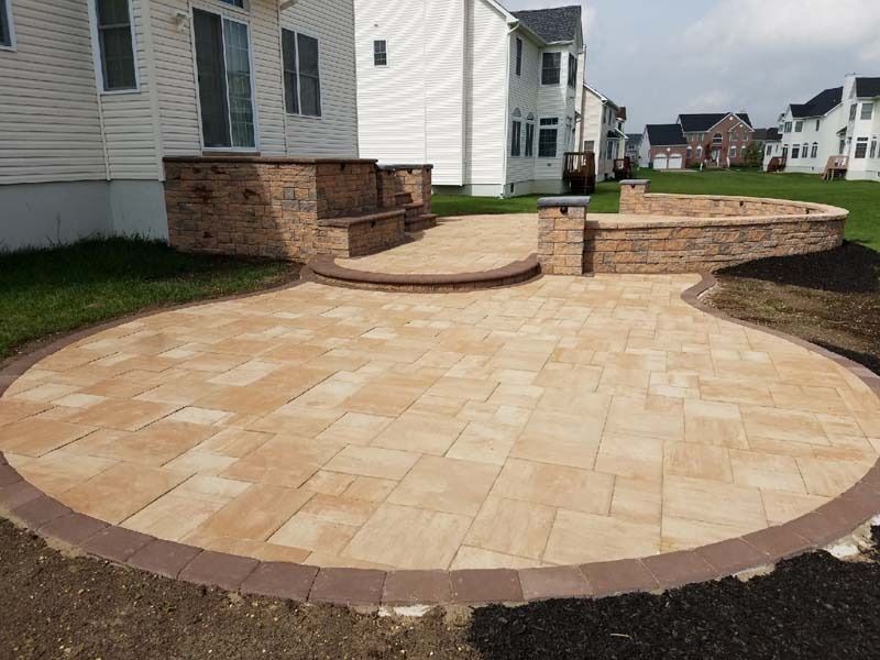 A paved patio with tan pavers, a curved wall, and steps, next to a house with a lawn.