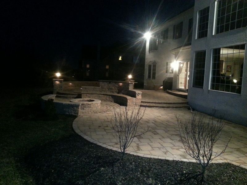 Nighttime backyard patio with fire pit, steps, and house with lit windows.