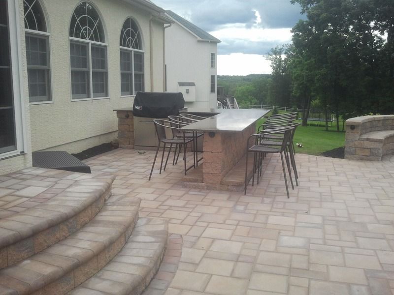 Patio with grill, large counter with bar stools, and steps. Brick pavers, beige tones, and outdoor setting.