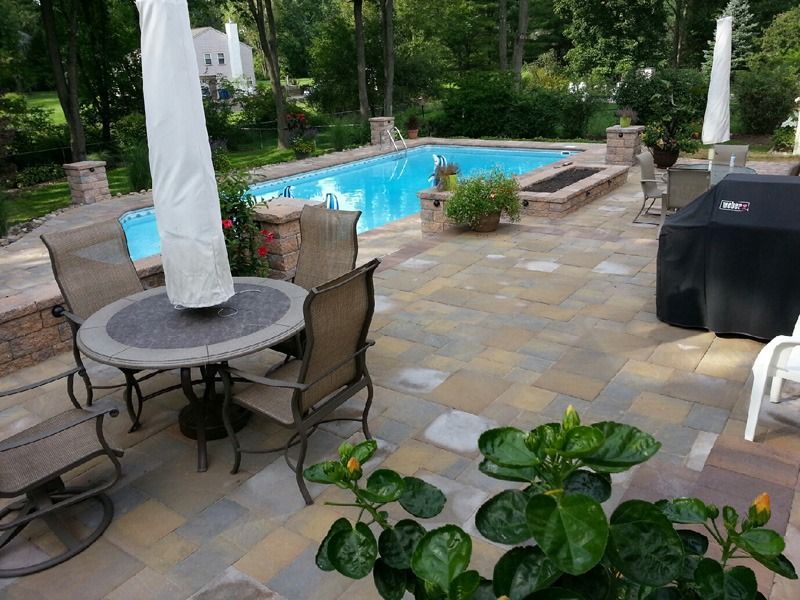 Patio with a pool, seating area, and grill. Blue pool, stone patio, green foliage.