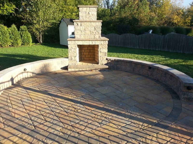 Outdoor patio with a stone fireplace, brick pavers, and a retaining wall, under a clear sky.