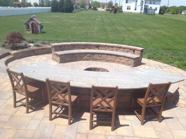 Circular patio with a built-in fire pit, stone seating, and wood chairs in a backyard.