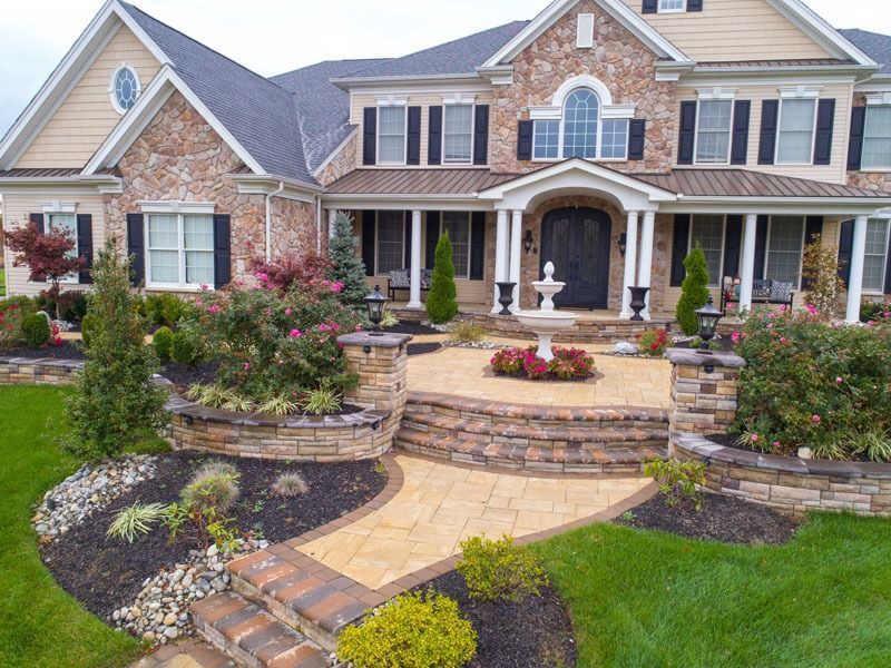 Stone-fronted house with a paved patio, fountain, and manicured landscaping, including flowers and shrubs.