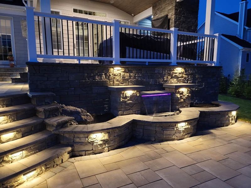 Outdoor patio with stone stairs, retaining walls, and a small waterfall illuminated by lights at night.