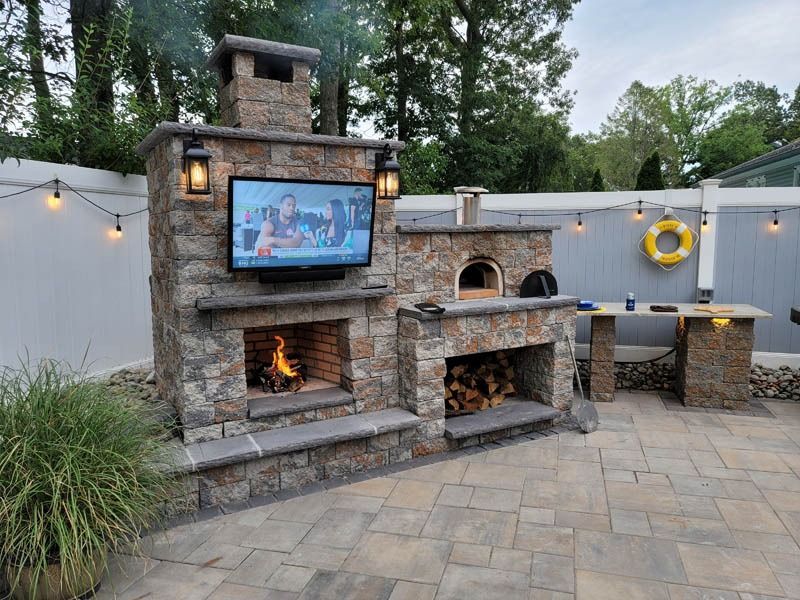 Outdoor stone fireplace with TV, pizza oven, and wood storage. Patio setting.