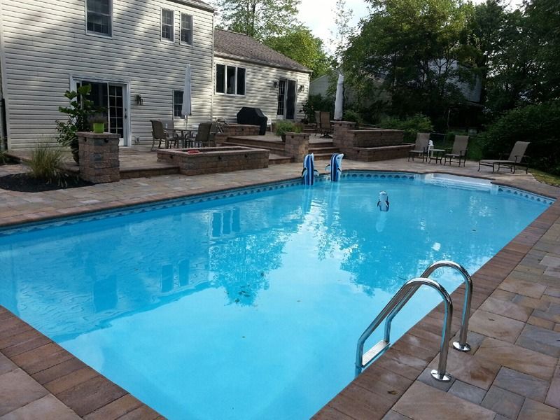 Backyard pool with tan pavers, steps, and lounge chairs. Beige house in the background. Blue water.