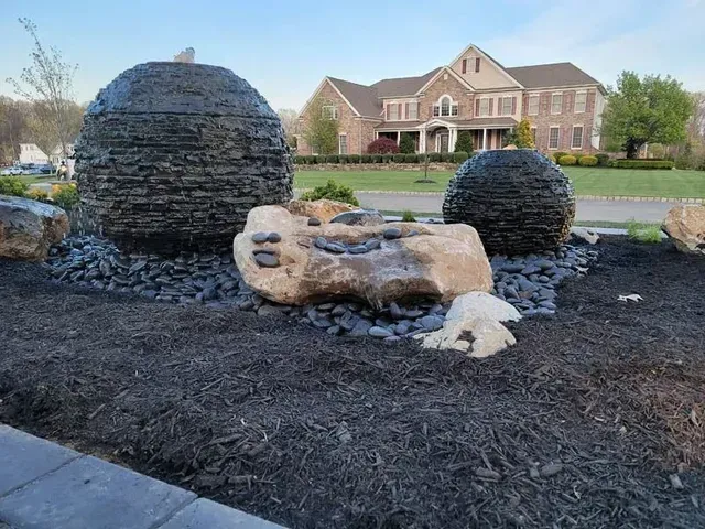 Two dark, textured stone sphere water fountains sit among light-colored landscape rocks, set before a large brick house.