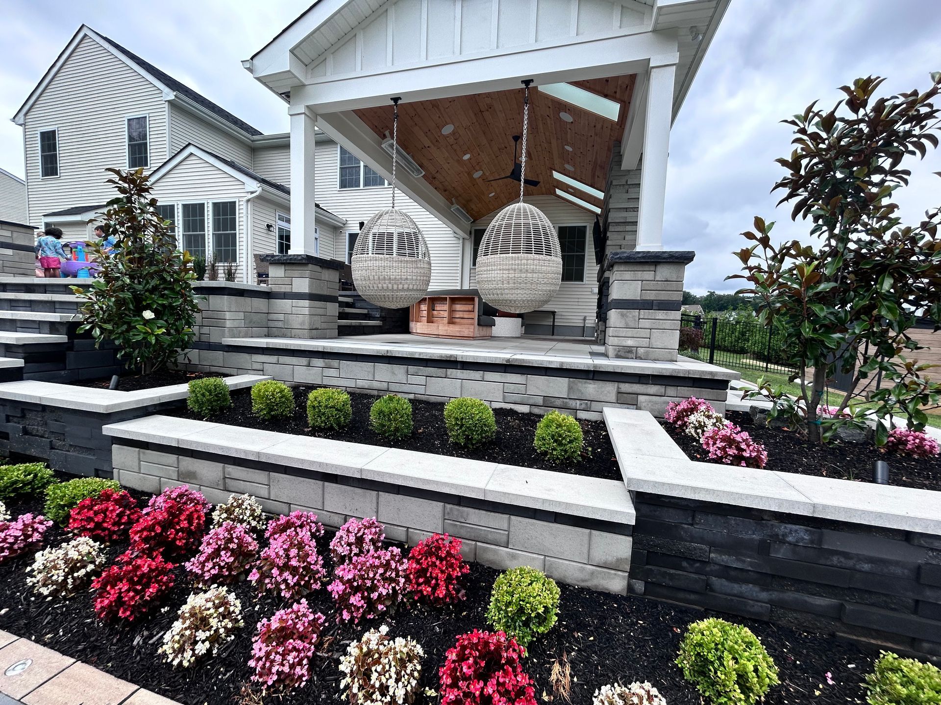 Landscaped patio with hanging egg chairs, tiered flower beds, and a white house.
