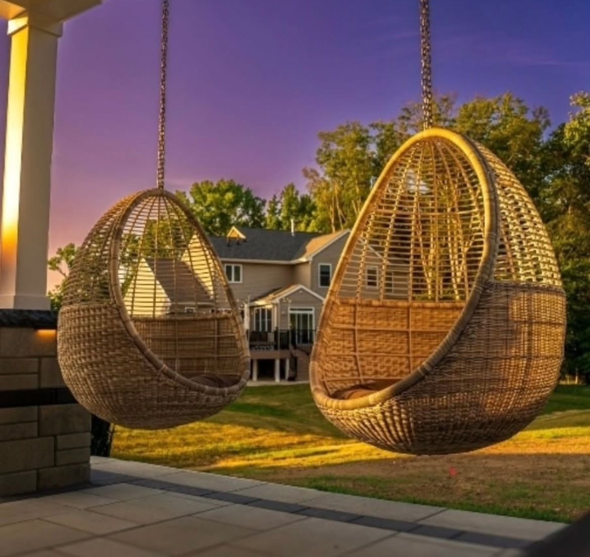 Two hanging wicker egg chairs on a patio overlooking a house at sunset with a purple sky.