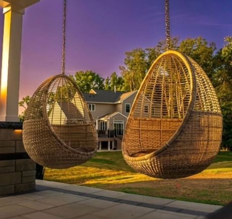 Two hanging wicker egg chairs on a patio overlooking a house at sunset with a purple sky.