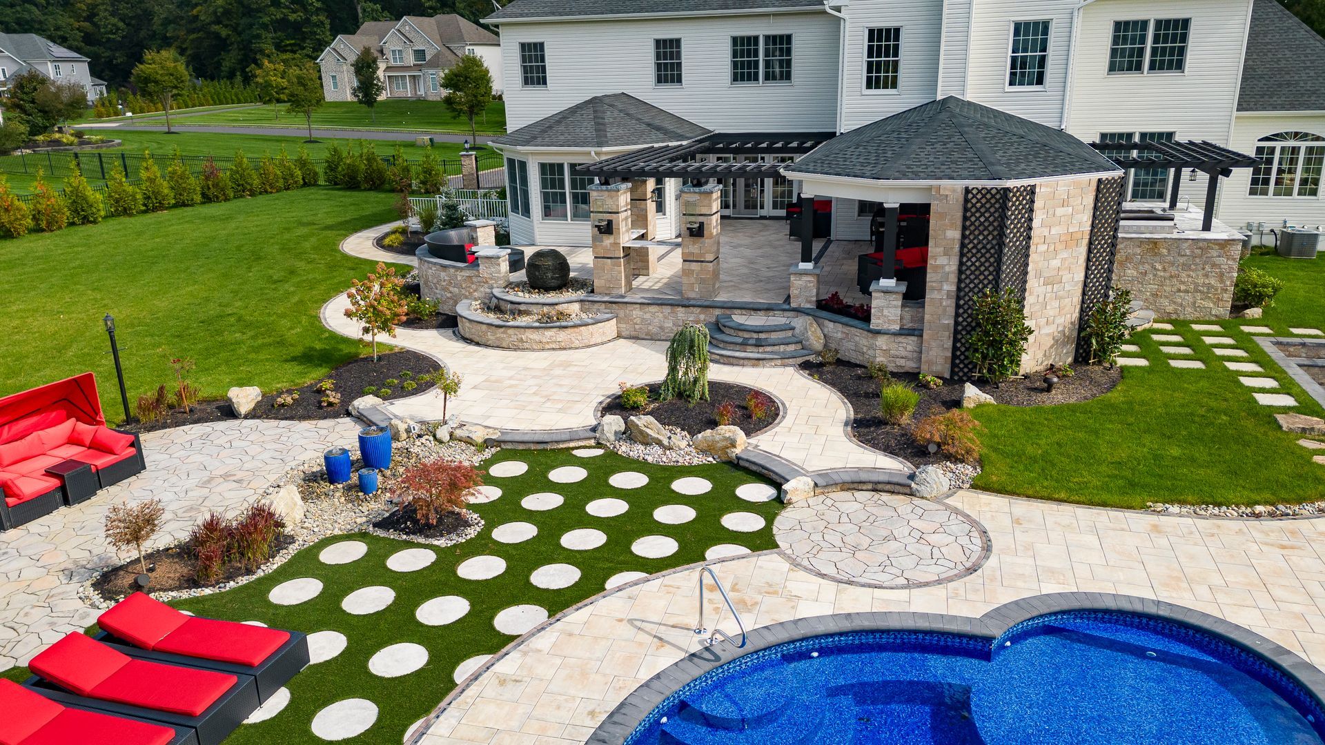 Aerial view of a luxury backyard with a pool, red lounge chairs, stone patio, green turf with circular pavers, and pergola.