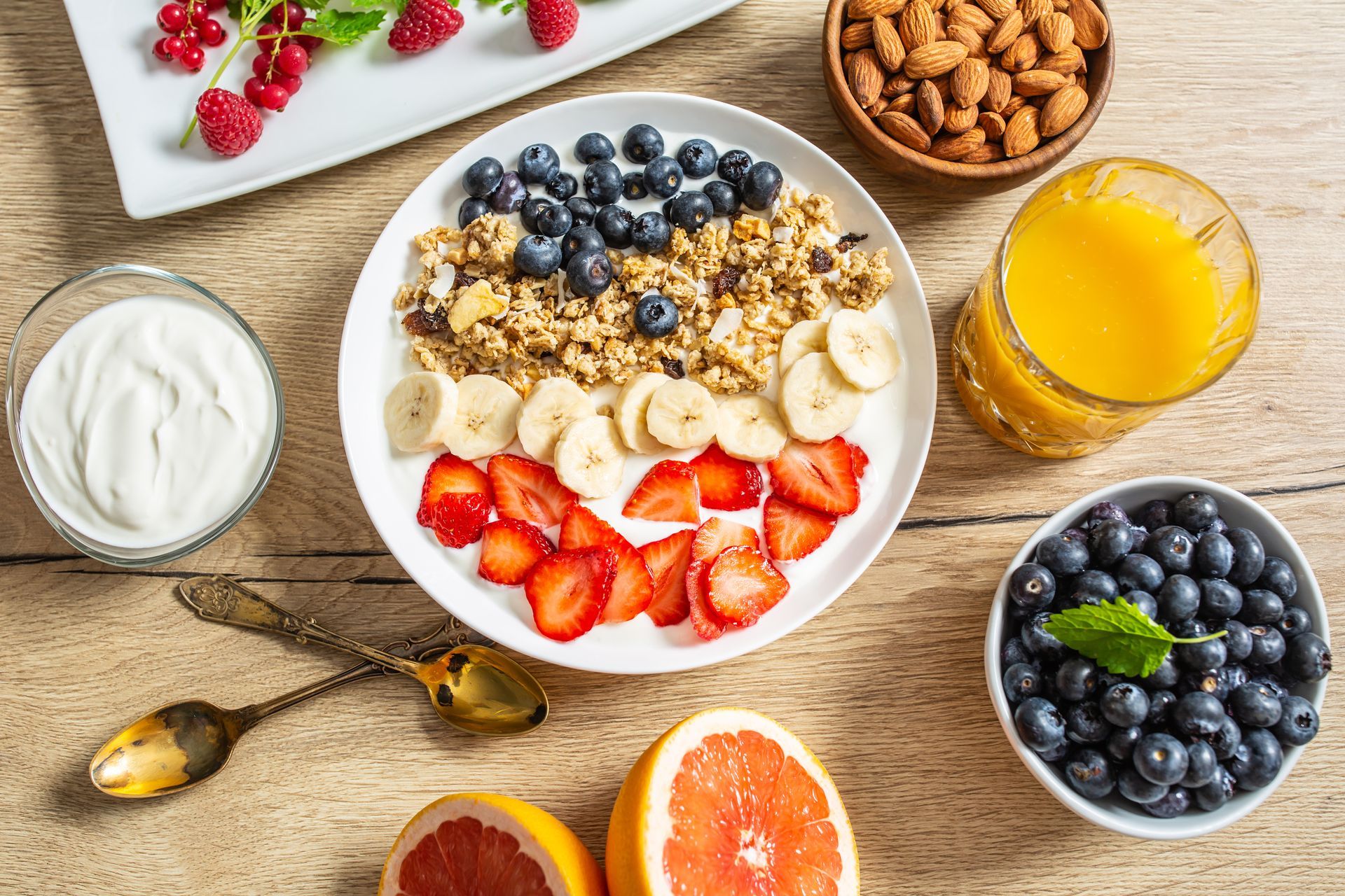 A bowl of granola with bananas , strawberries , blueberries , almonds and yogurt on a wooden table.