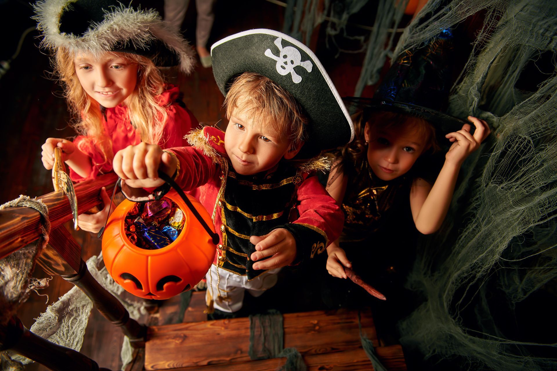 Three children in Halloween costumes: pirate, pirate, and witch, holding a pumpkin candy bucket.
