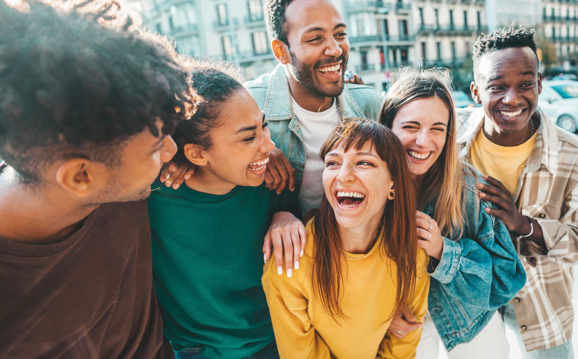 Group of smiling friends, arms around each other, laughing outdoors in front of a building.