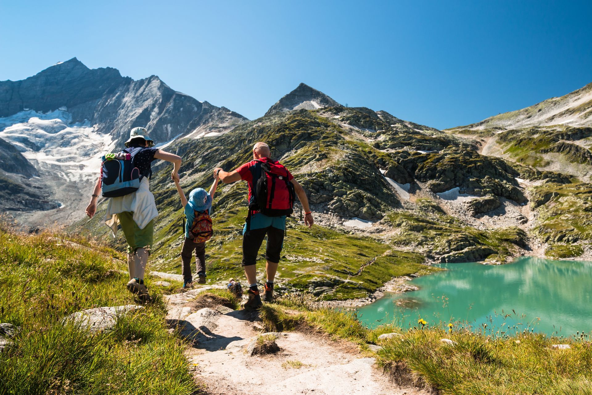 Family hiking along a trail near a turquoise lake, mountains in the background on a sunny day.