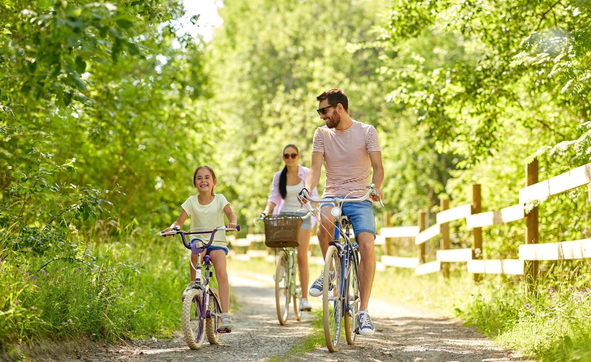 Family of three riding bikes on a sunny path through a green wooded area.
