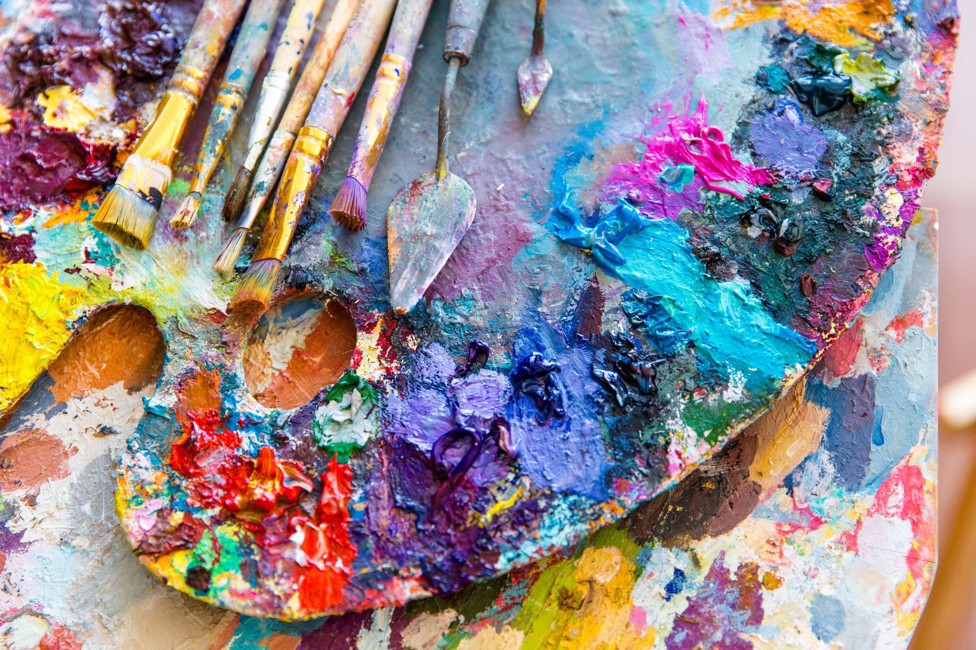 Palette with smears of colorful paint, paintbrushes, and palette knives.