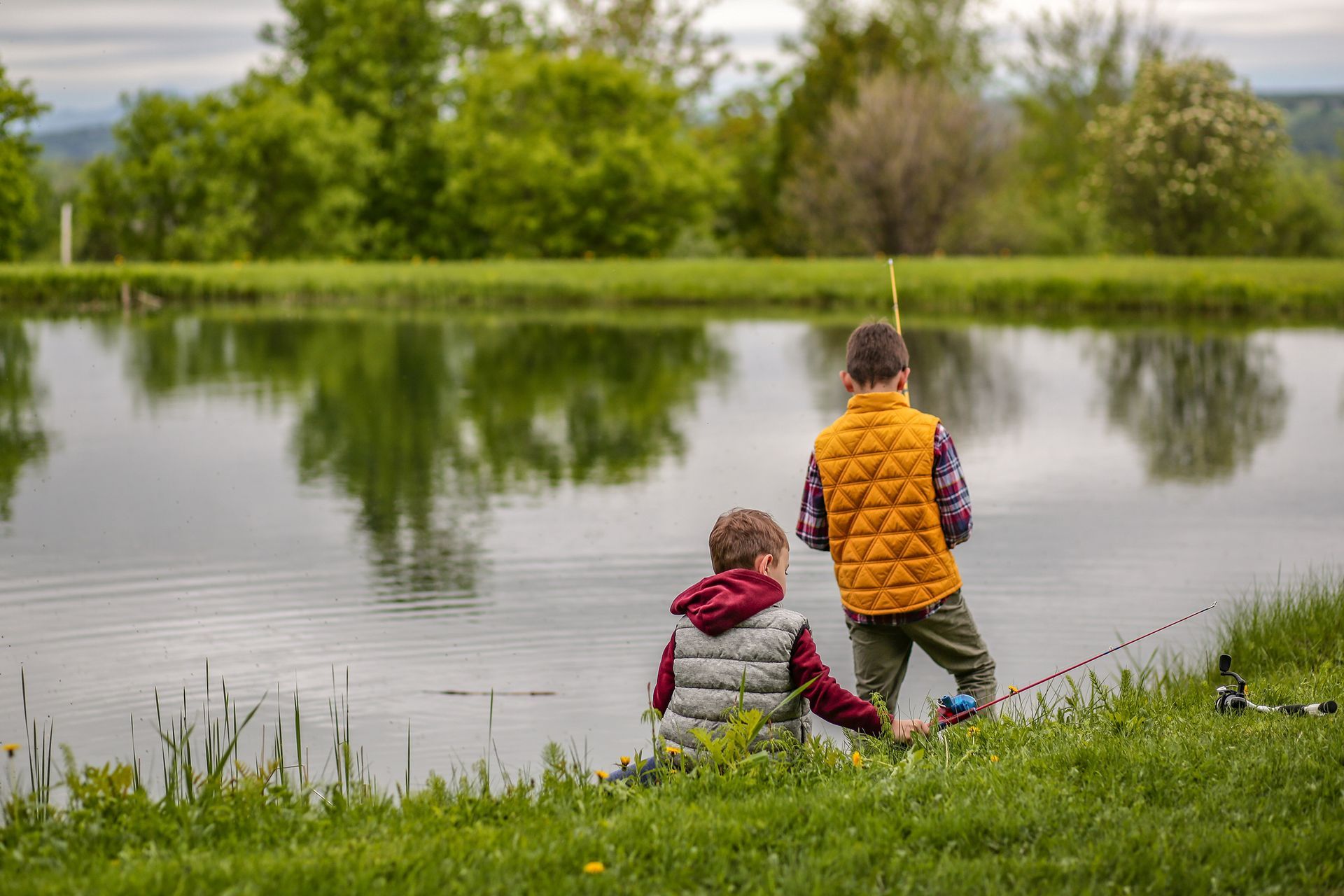 Two children sitting by a pond, one in an orange vest and one in a red jacket, with trees reflected in the water.