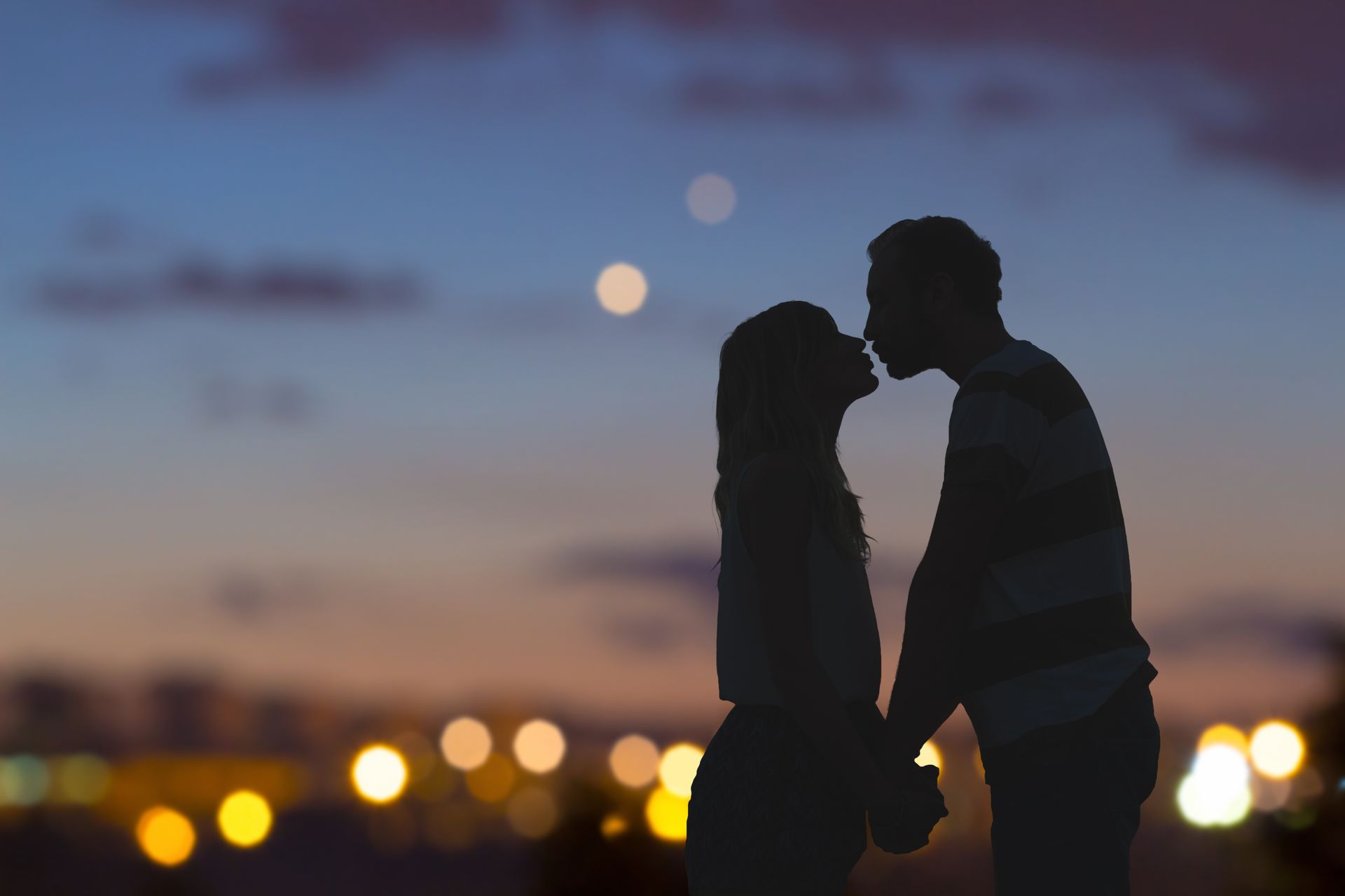 Silhouetted couple kissing, holding hands, at dusk with blurred lights.