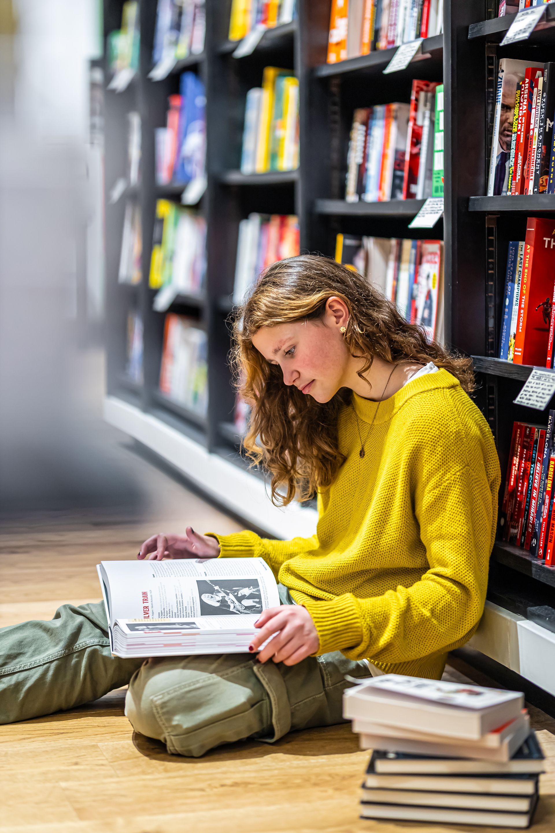 A person with curly hair wearing a yellow sweater sits on a bookstore floor reading, with a stack of books nearby.