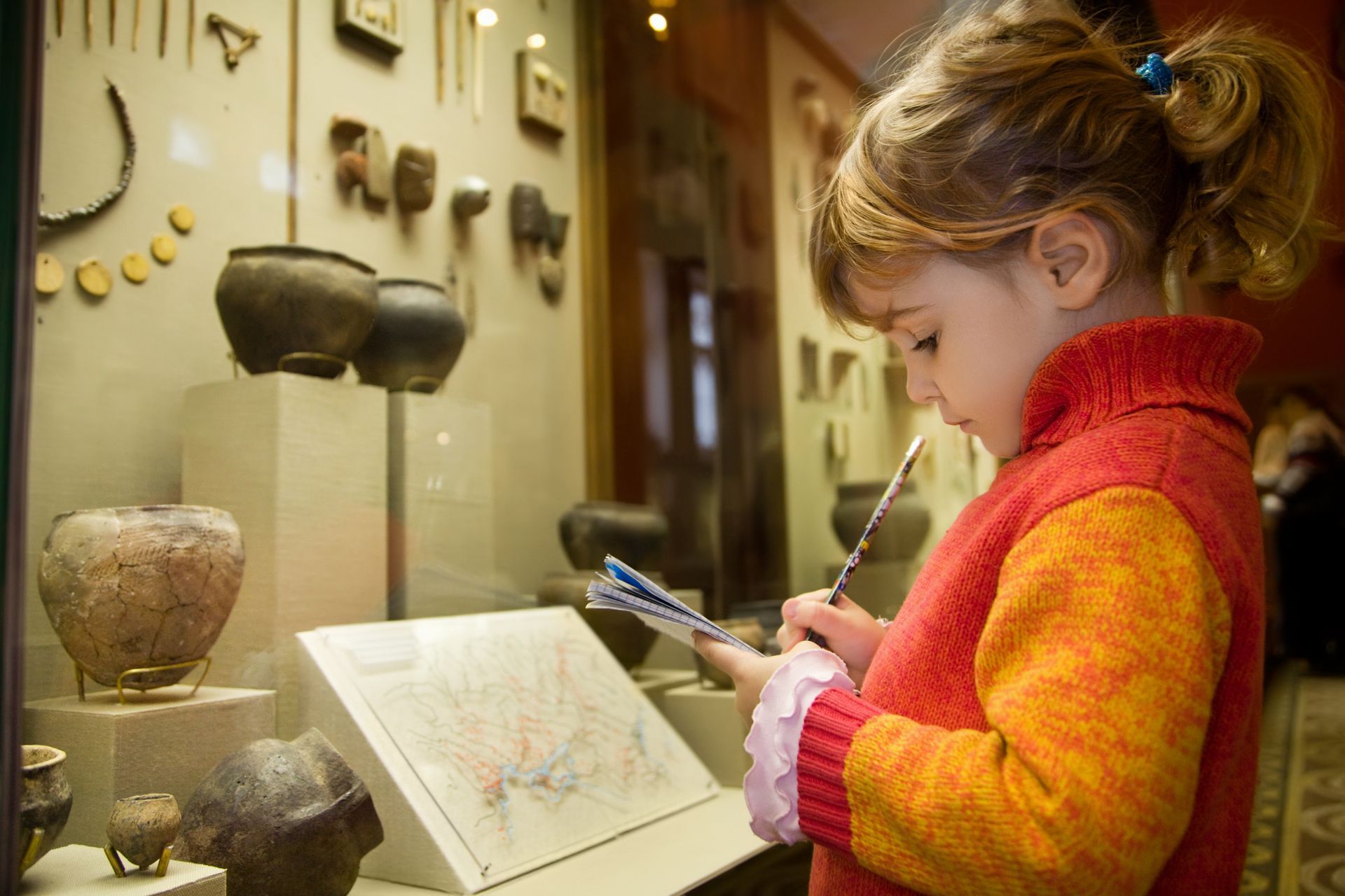 Girl sketching artifacts in a museum display case. Wearing an orange sweater, she holds a pencil and notebook.