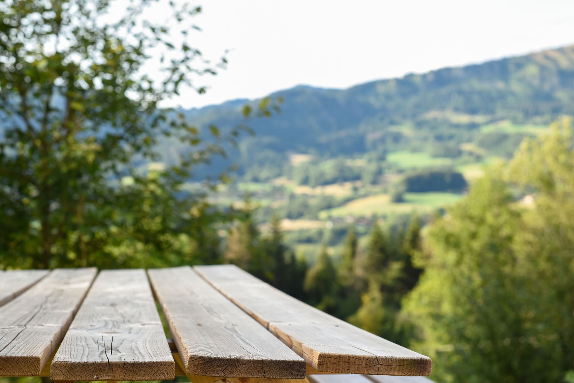 Wooden picnic table in the foreground overlooking a blurred, sunny mountain landscape.