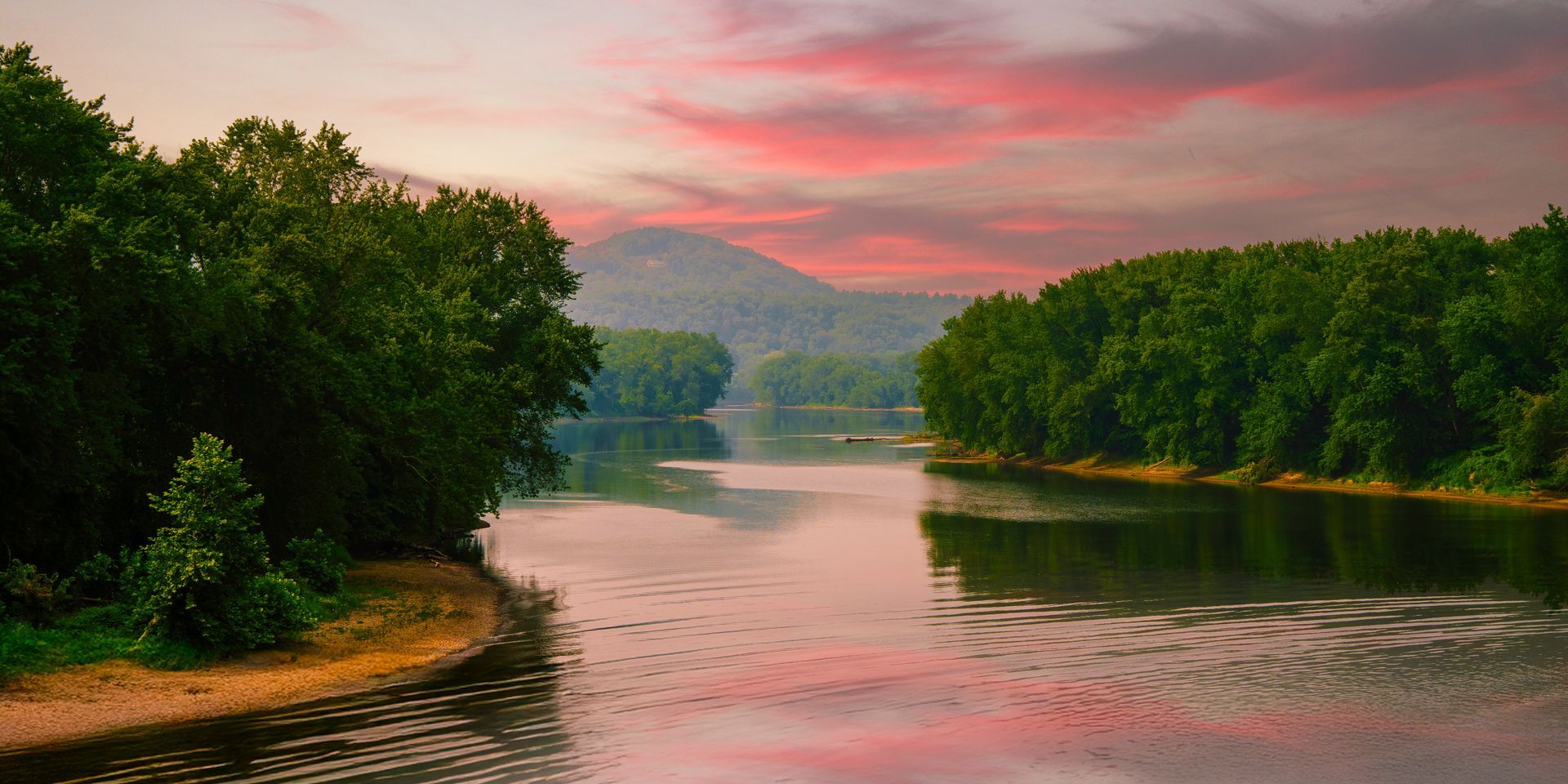 A tranquil river flows between lush green banks toward a distant mountain under a vibrant pink and orange sunset sky.