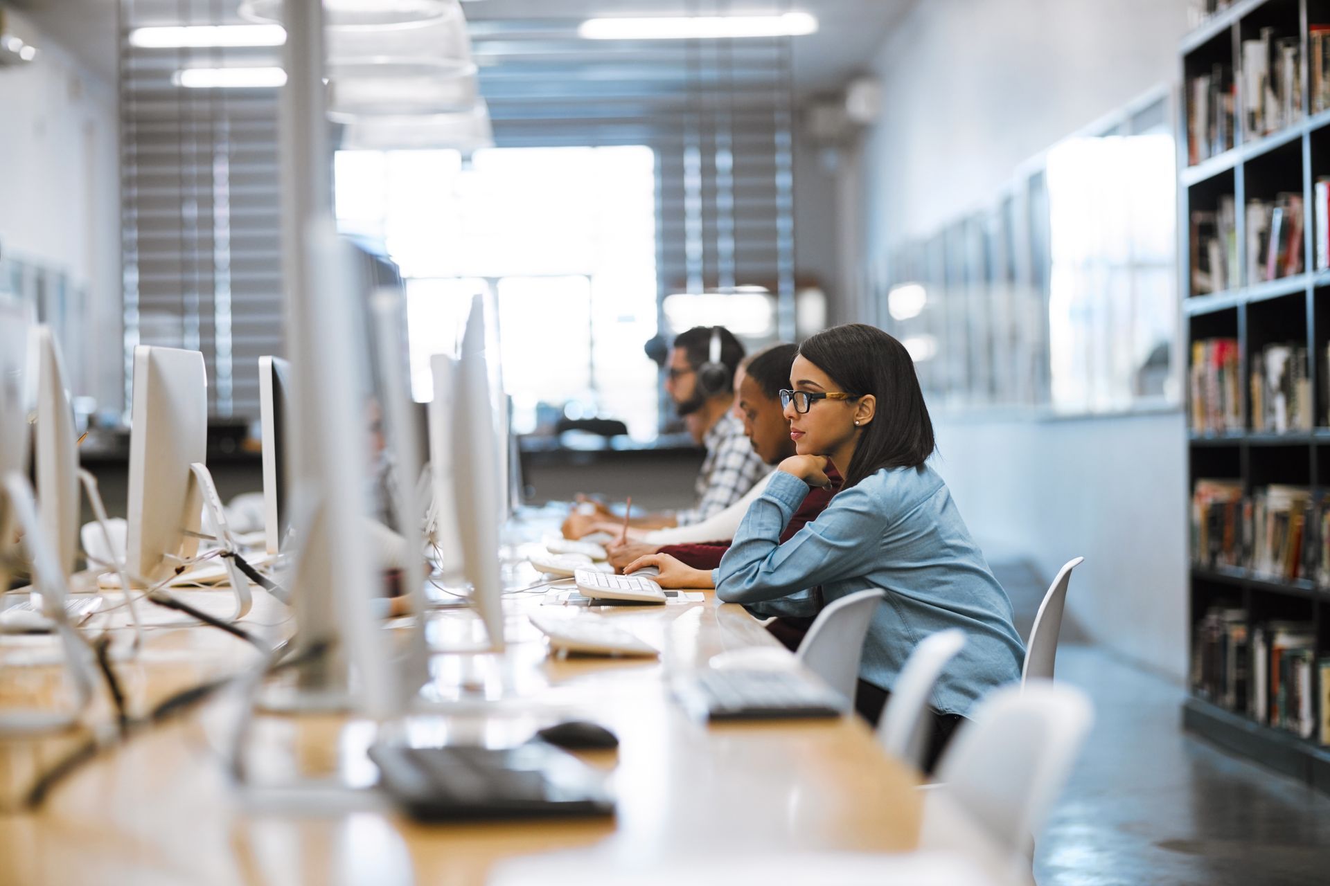 People sit at a row of computers in a library, focusing on their screens in a brightly lit, professional workspace.