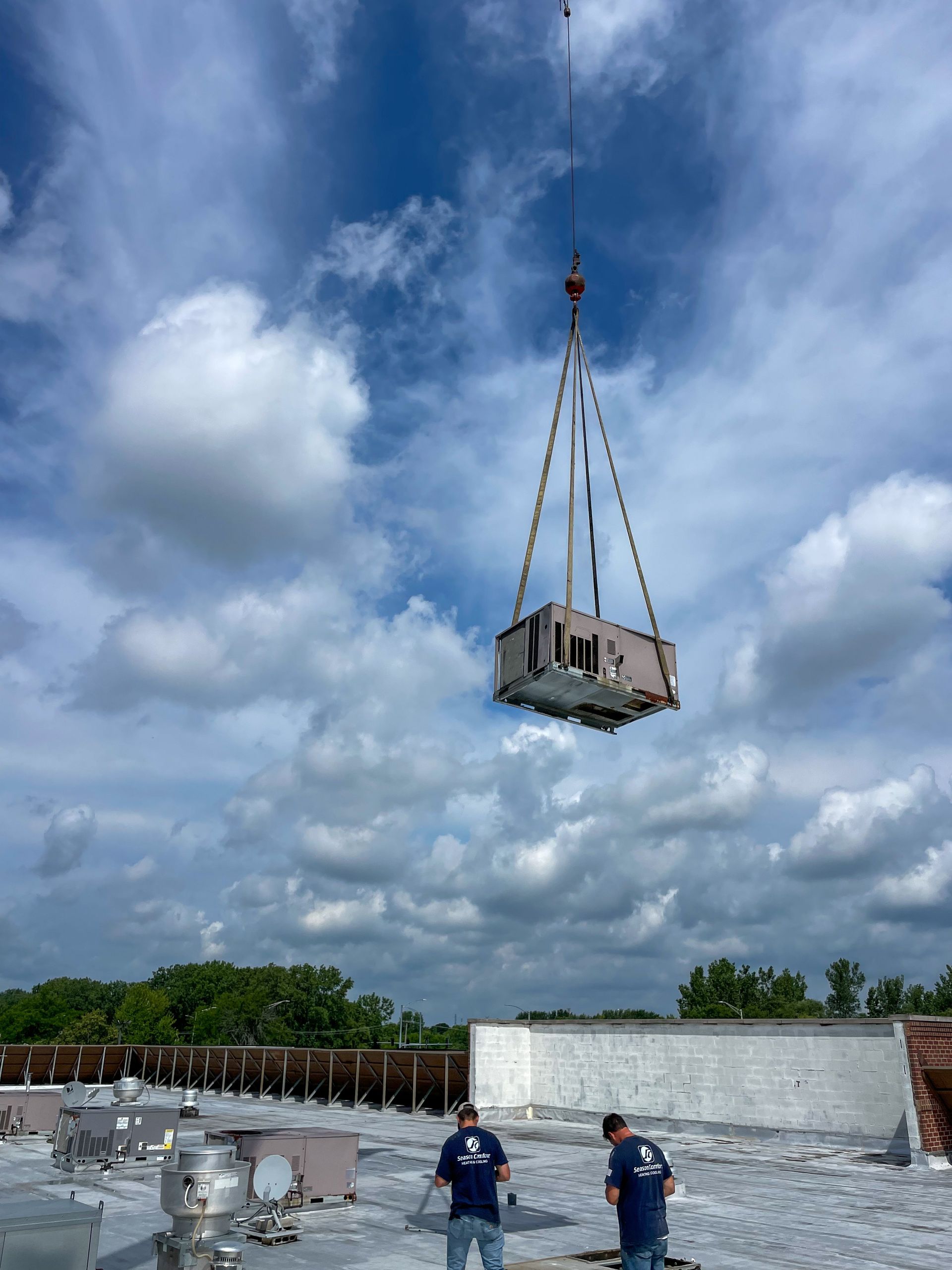 Crane lifting an HVAC unit onto a commercial building rooftop. Two workers in blue shirts observe. Cloudy sky.