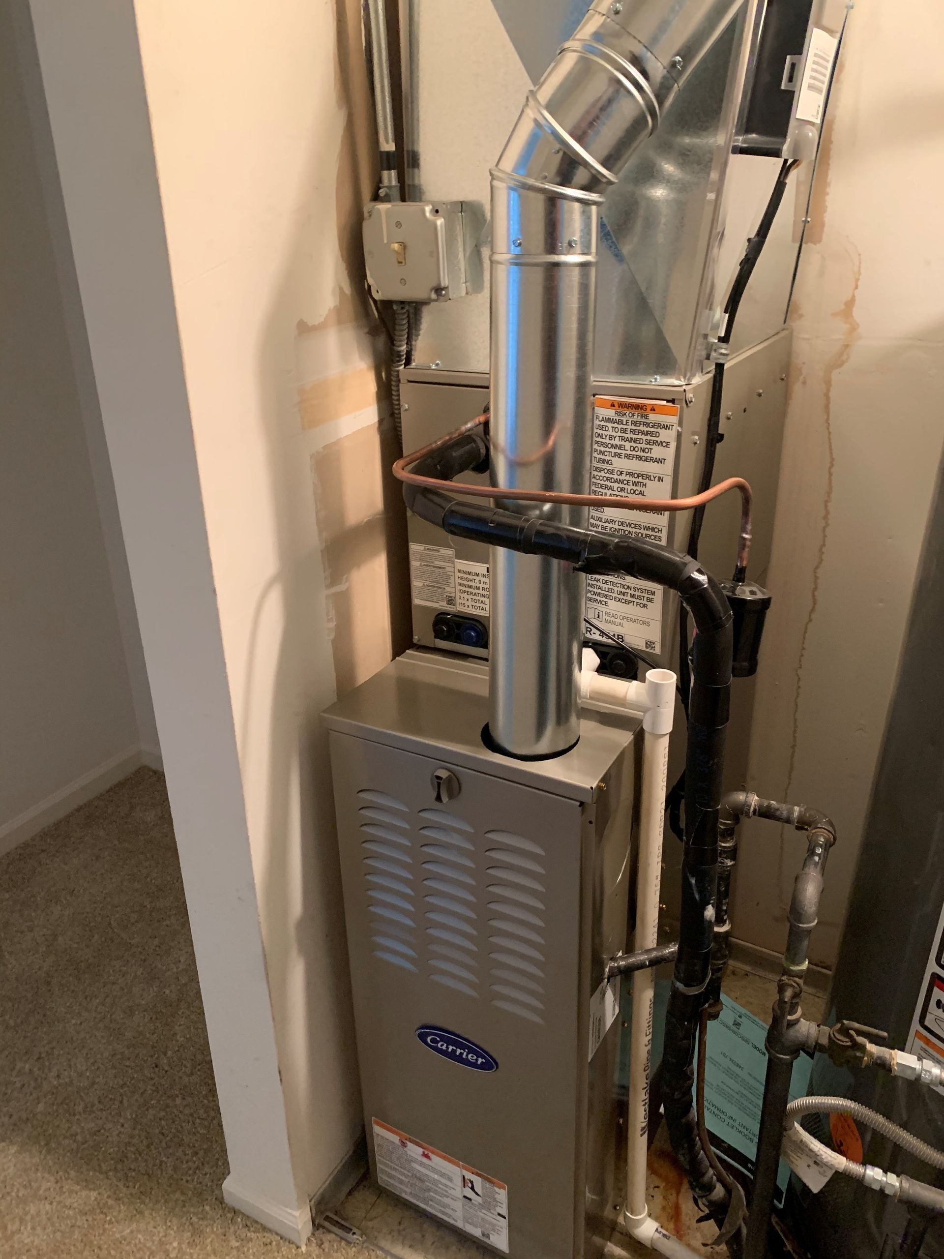 A Carrier furnace in a small closet, with silver vent pipes and various connections.