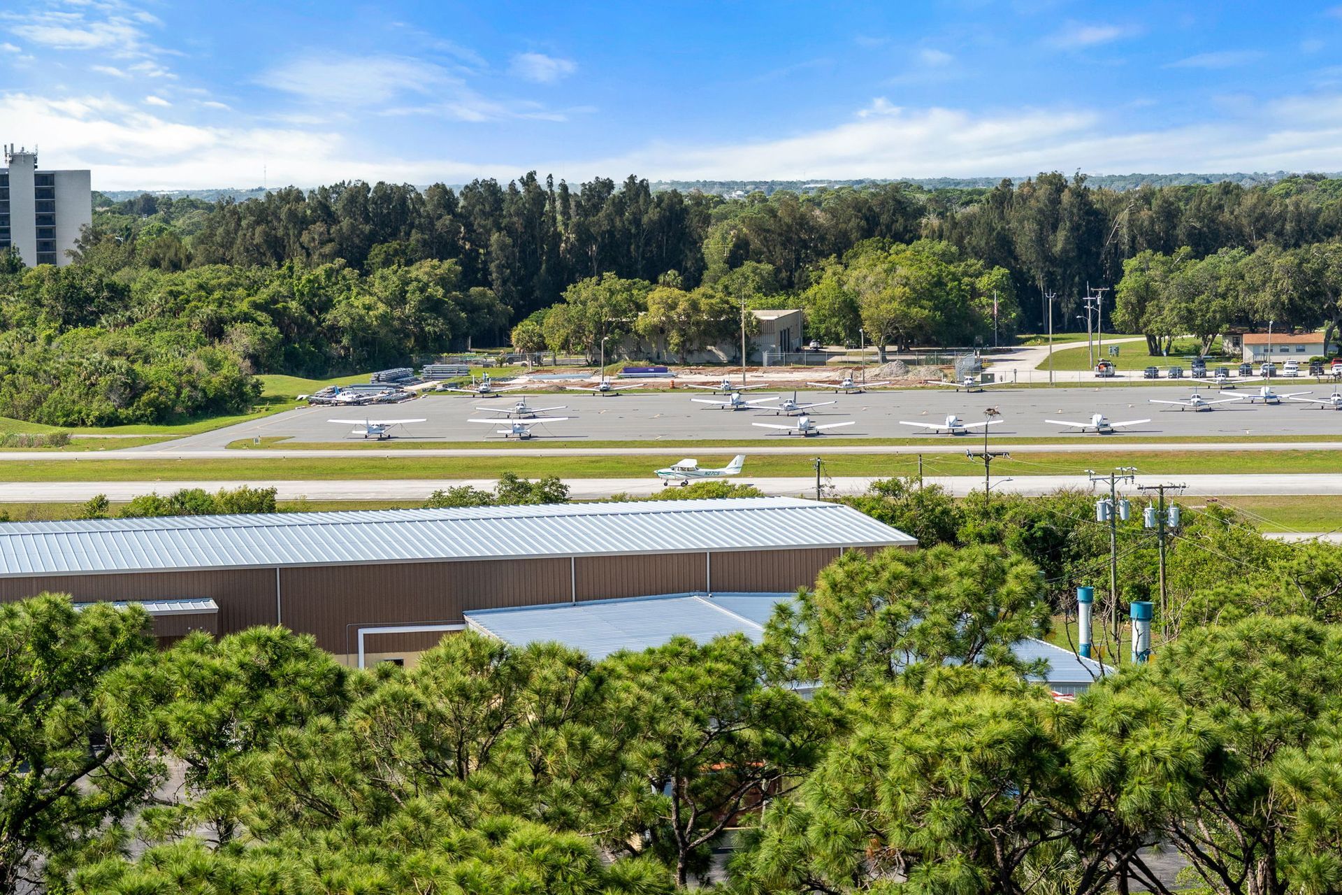 Wide outdoor view of a tree-lined road and parking lot beside a low building under a bright sky. View of private airplane hangers next to Merritt Island Airport