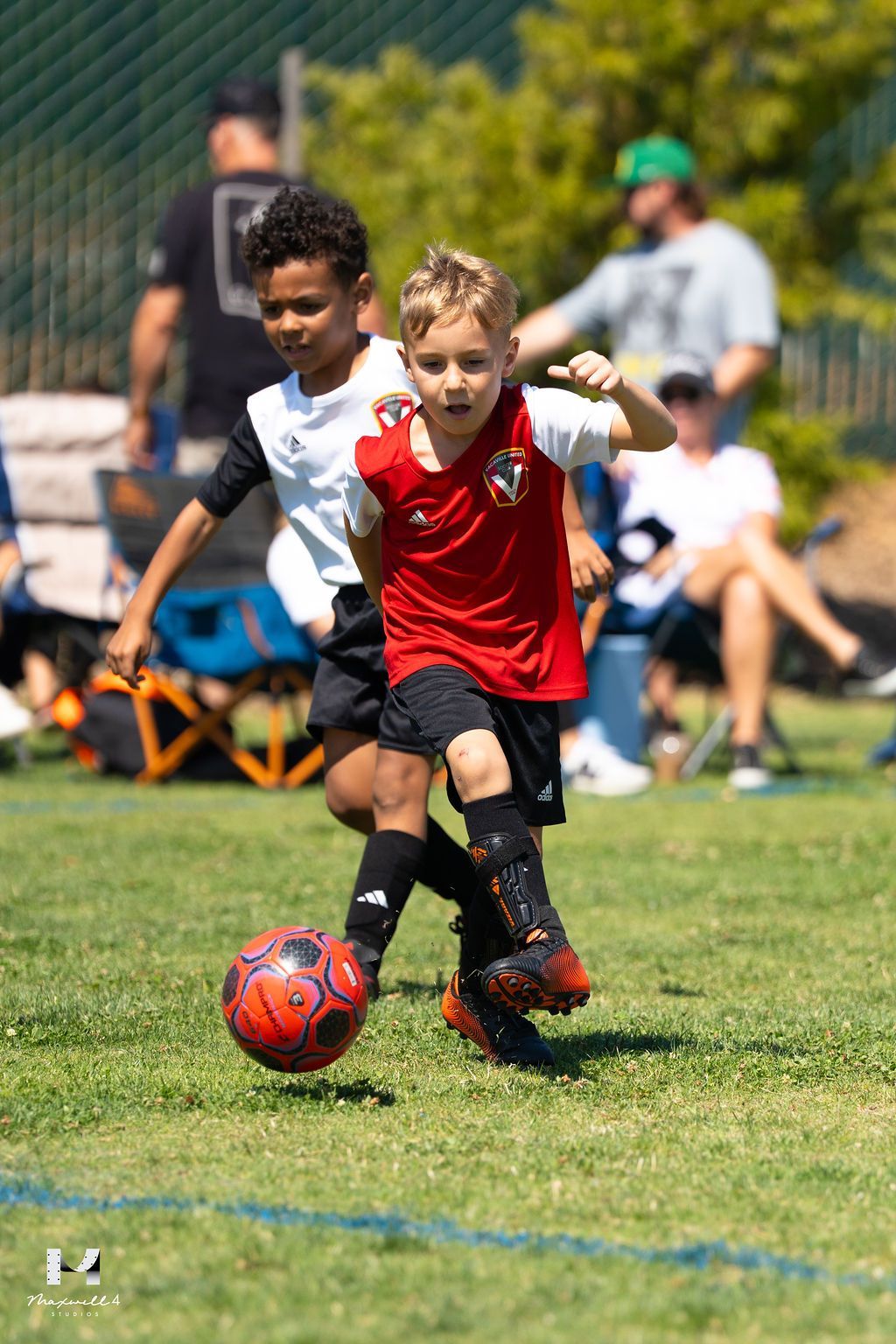 Two young soccer players on a green field. One in red kicks the ball, the other in white pursues.