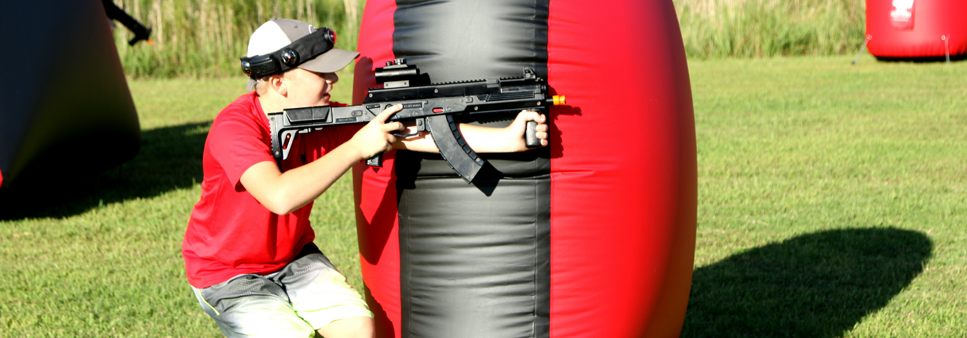 Person in a red shirt aiming a paintball gun from behind a red-and-black inflatable barrier on a grassy field