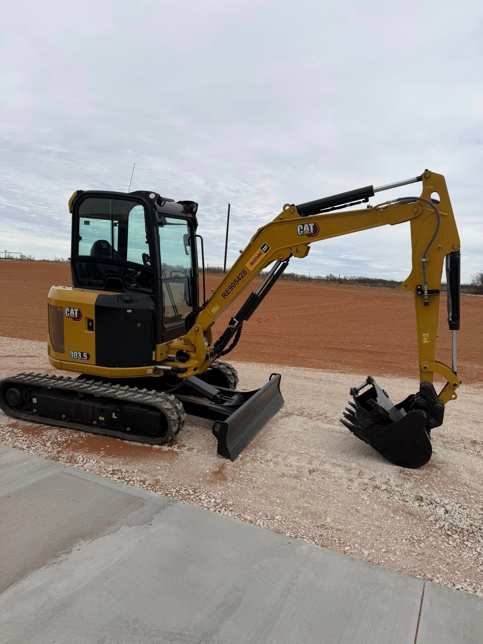 Yellow and black mini excavator on gravel, arm extended, in a field under a cloudy sky.