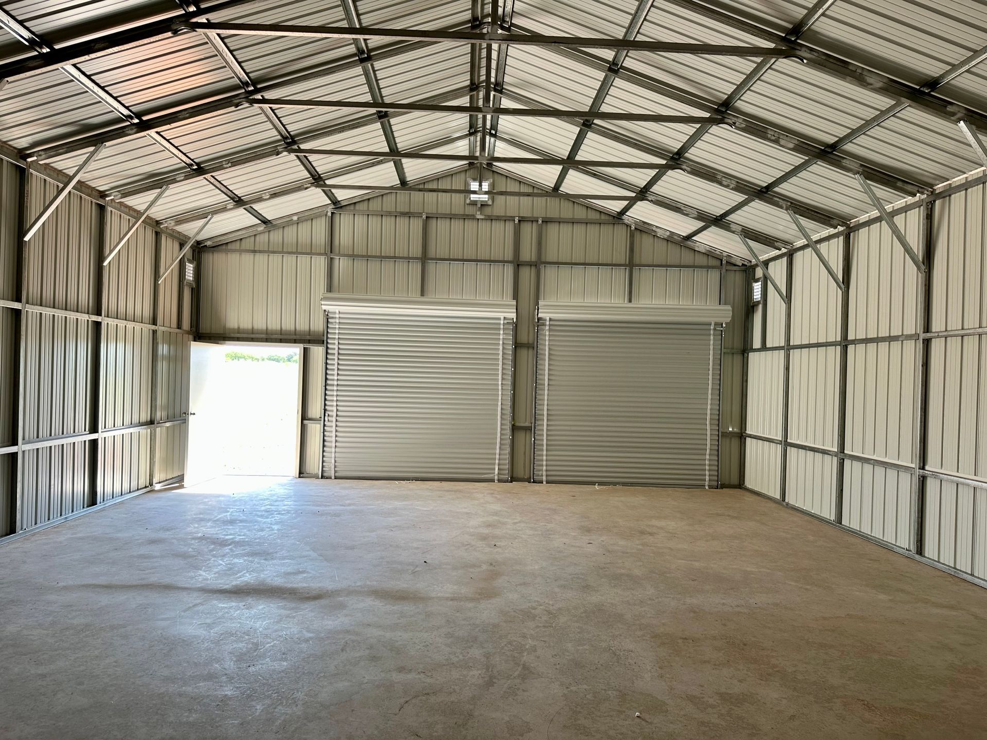 Inside of a metal storage shed with roll-up doors, bare concrete floor, and an open doorway.