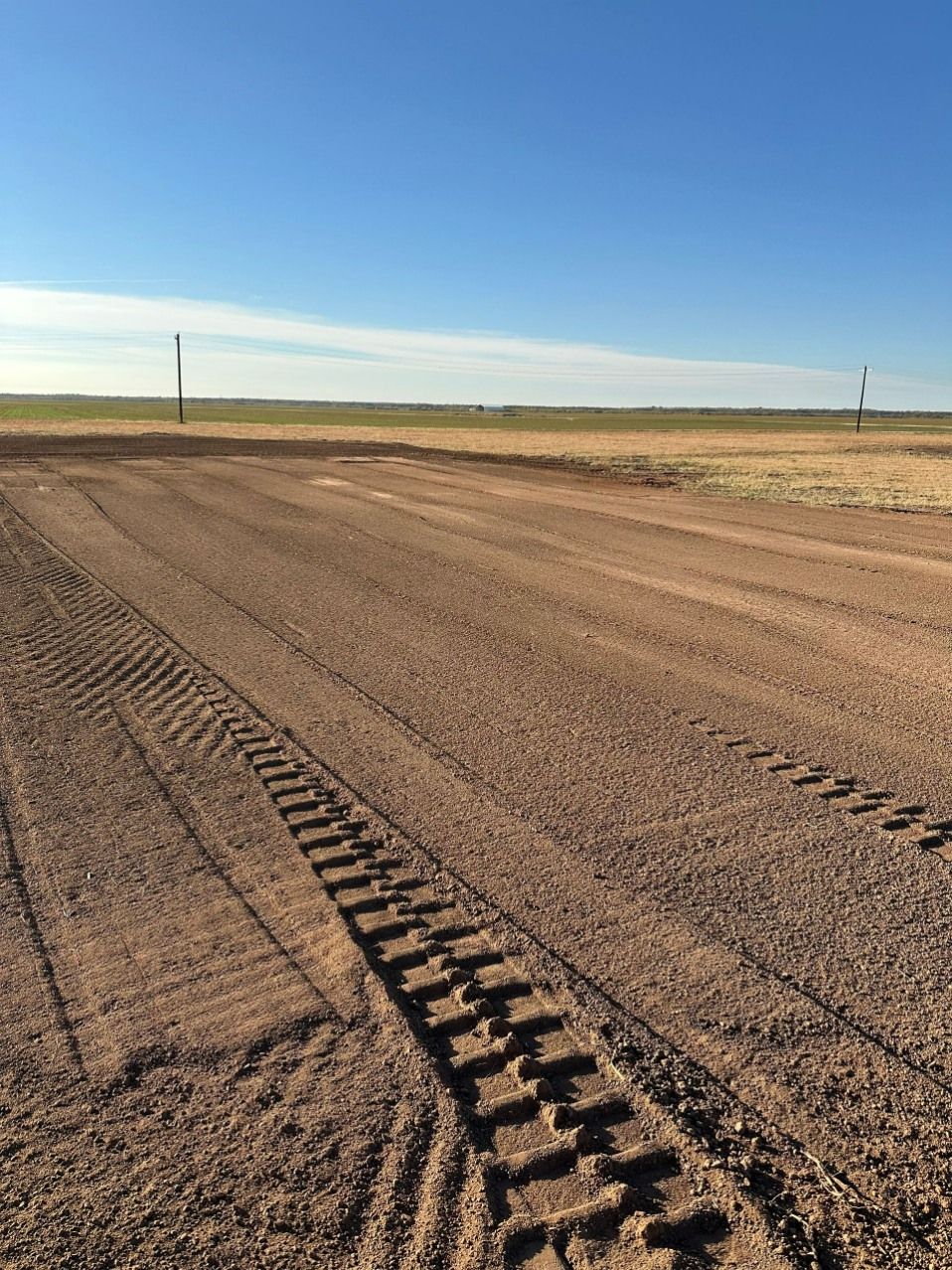 Dirt road with tractor tire tracks under a blue sky, leading to a flat, grassy horizon.