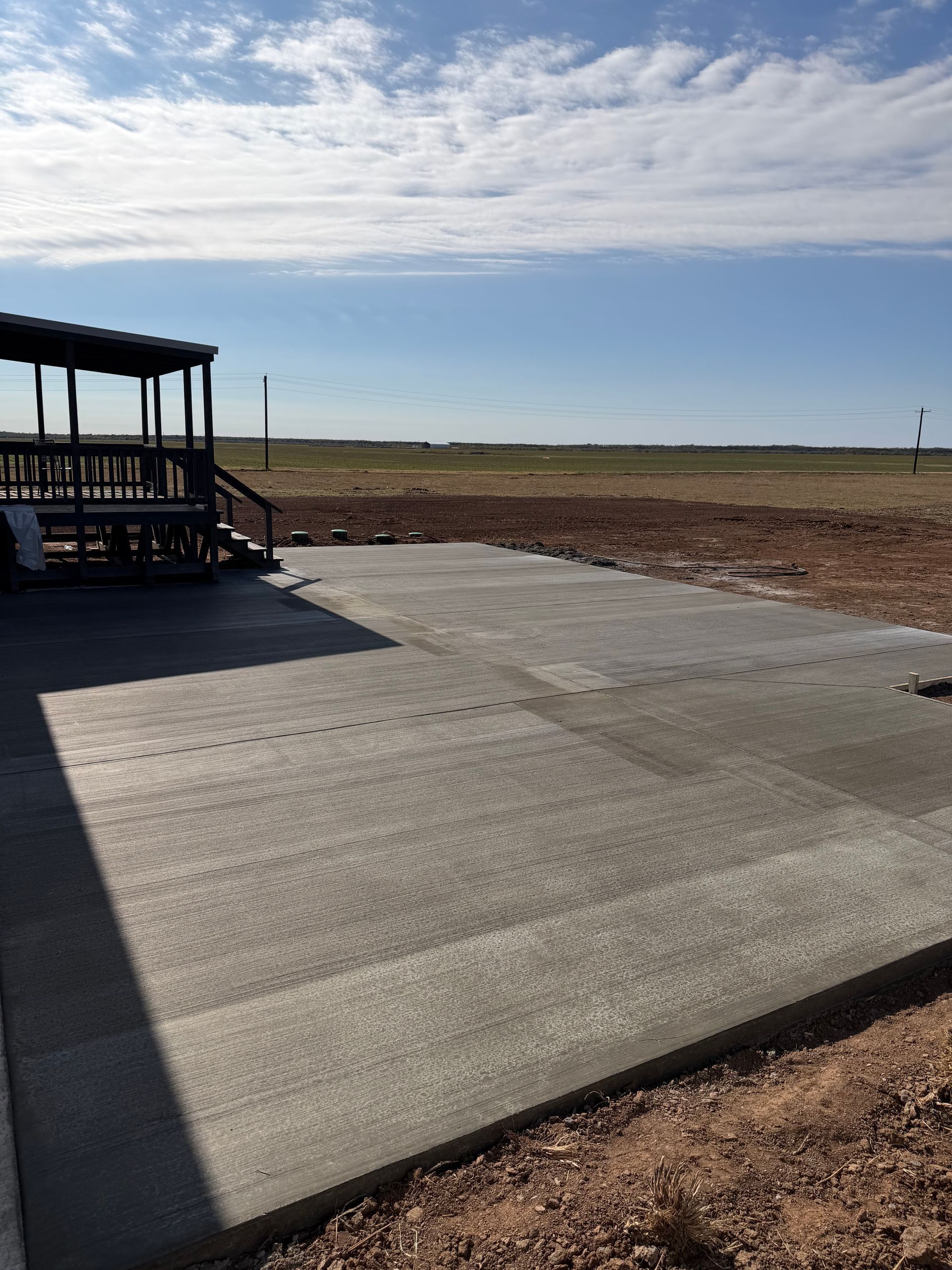 Newly poured concrete patio next to a wooden deck, with a field and blue sky in the background.
