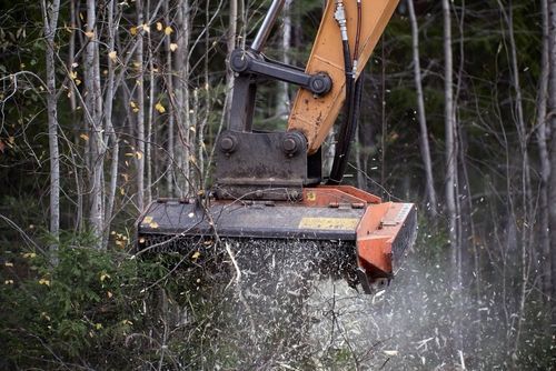 An excavator's brush cutter mulching trees in a forest; wood chips flying.