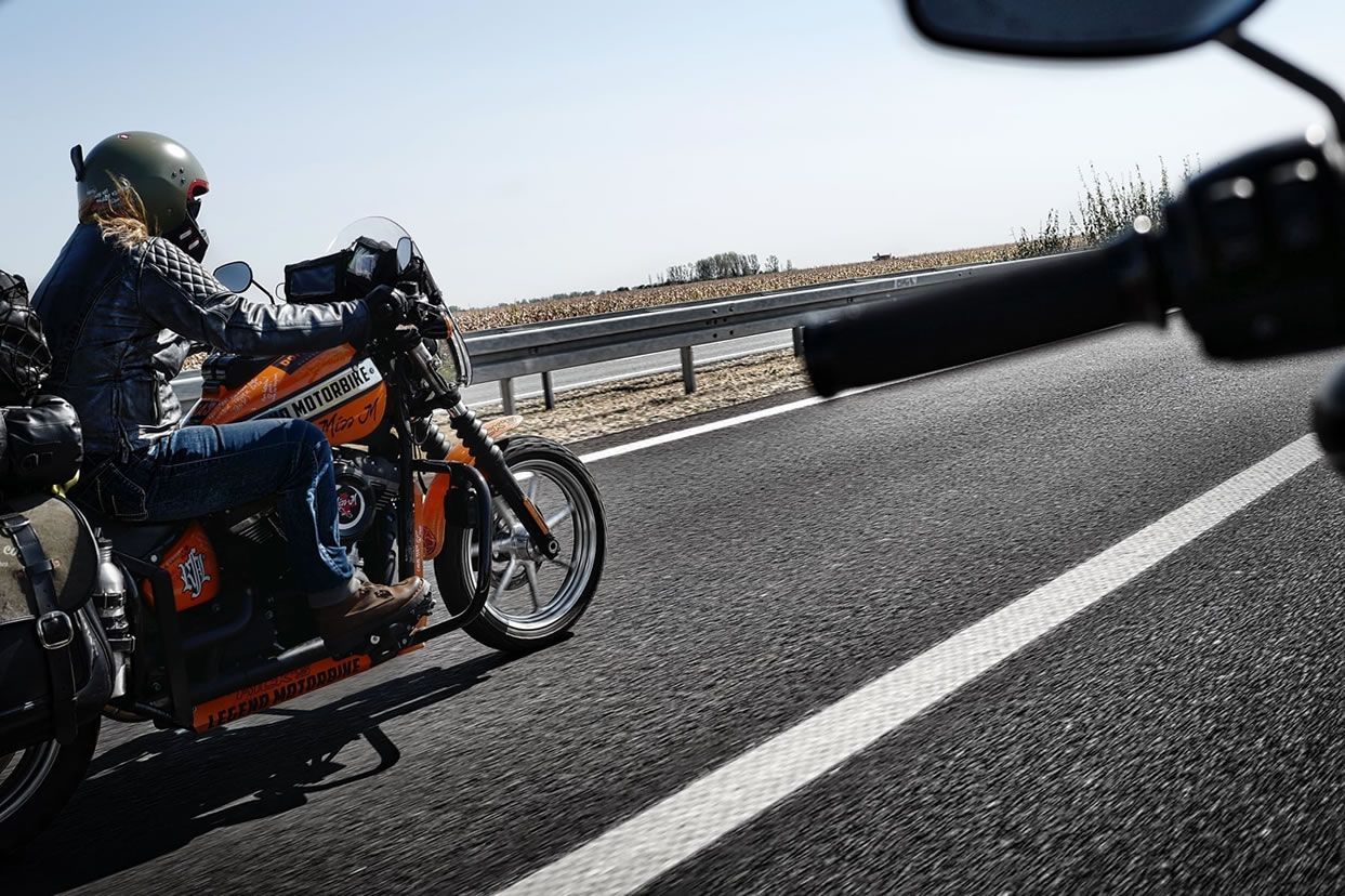 Un motocycliste sur un vélo orange circulant sur une route avec des champs ouverts par une journée ensoleillée.