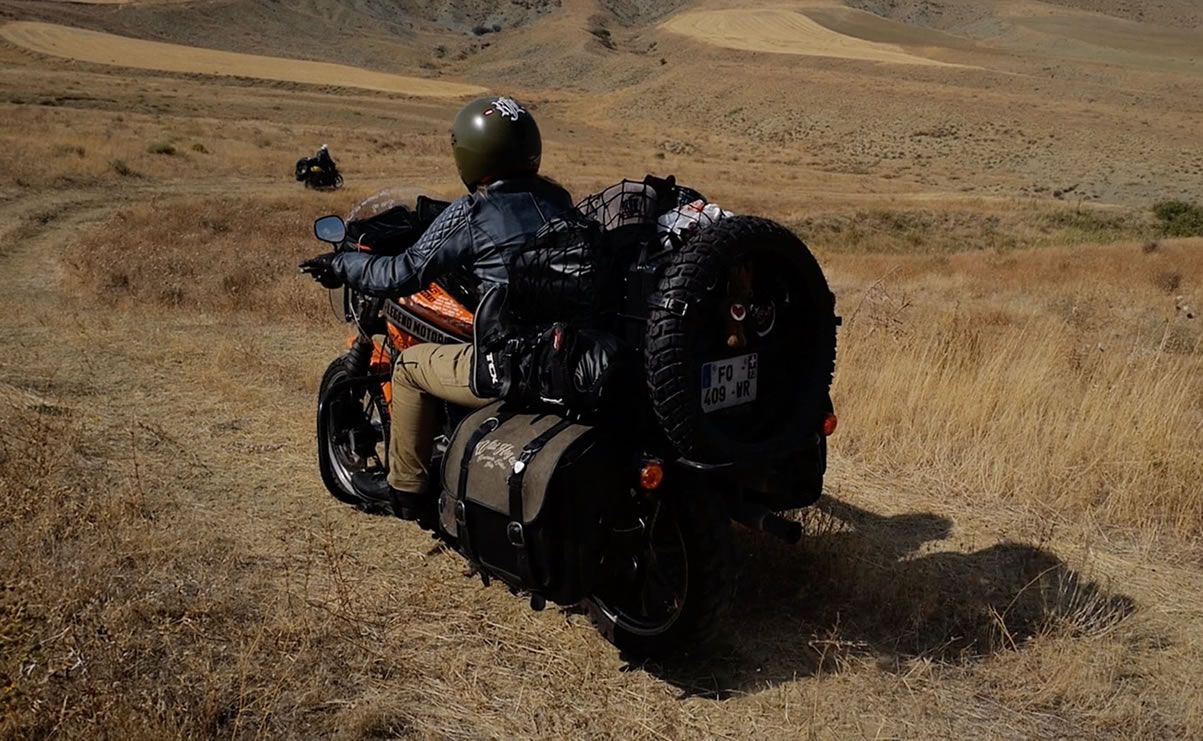 Une personne conduisant une moto lourdement chargée d'équipement à travers un paysage sec et ouvert.