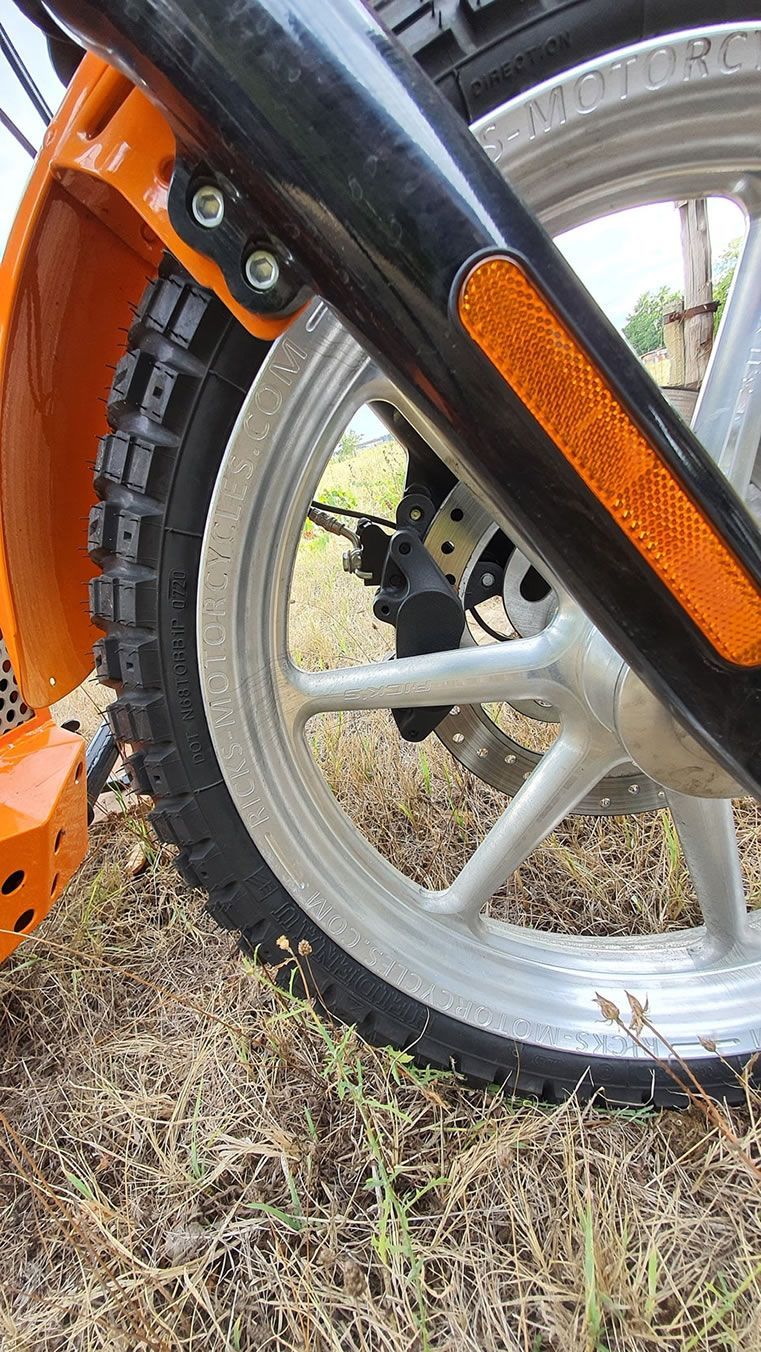 Gros plan d'une roue avant de moto orange et noire avec une jante argentée et un pneu noir sur de l'herbe sèche.