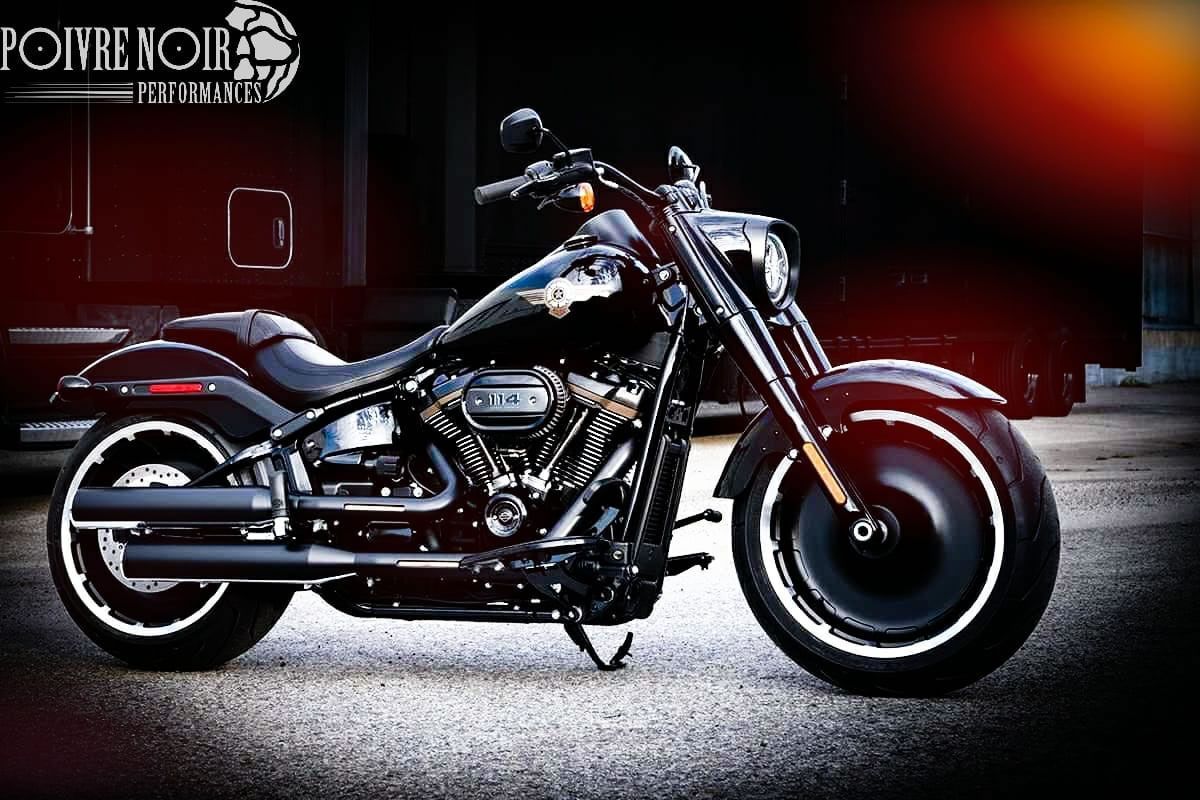 Moto Harley-Davidson noire, garée en extérieur. Caractéristiques : pneus larges, peinture foncée et phare rond.