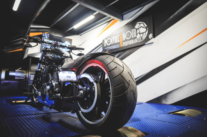 Moto sur un banc d'essai dans un garage, noire et chromée, avec logo sur le mur.