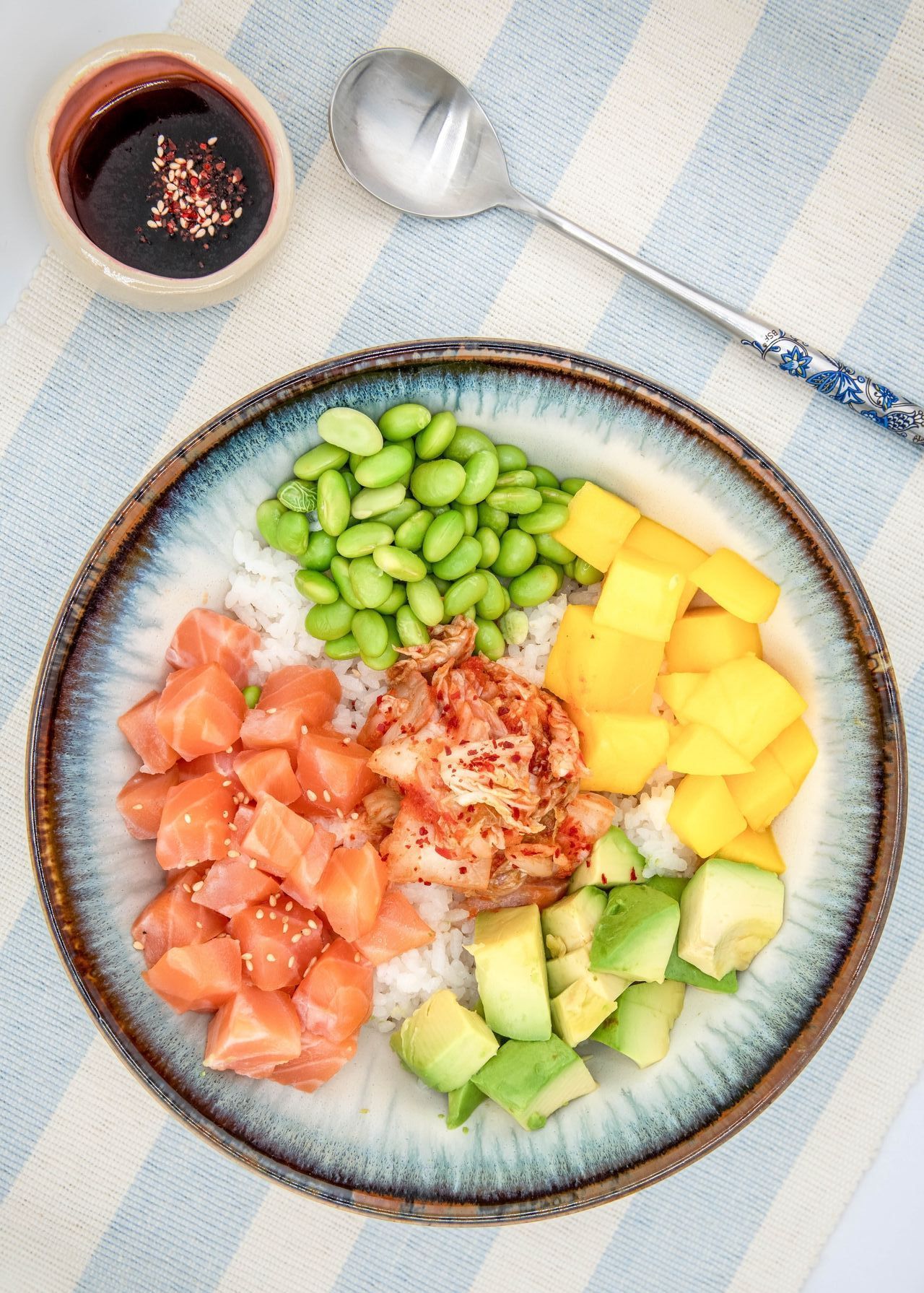 Poke bowl avec saumon, avocat, mangue, edamame et kimchi, avec sauce soja et cuillère.
