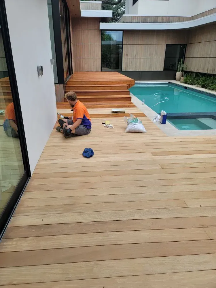 Person working on a wooden deck by a pool. Orange and blue shirt, seated on the deck.