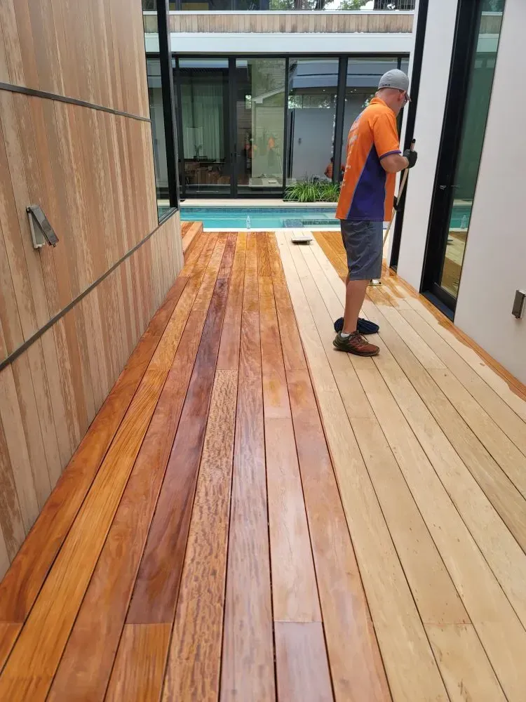 A person applying stain to a wooden deck next to a building with a pool.
