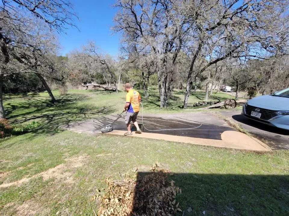 Person using a blower to clear leaves off a concrete path in a grassy area with trees under a blue sky.