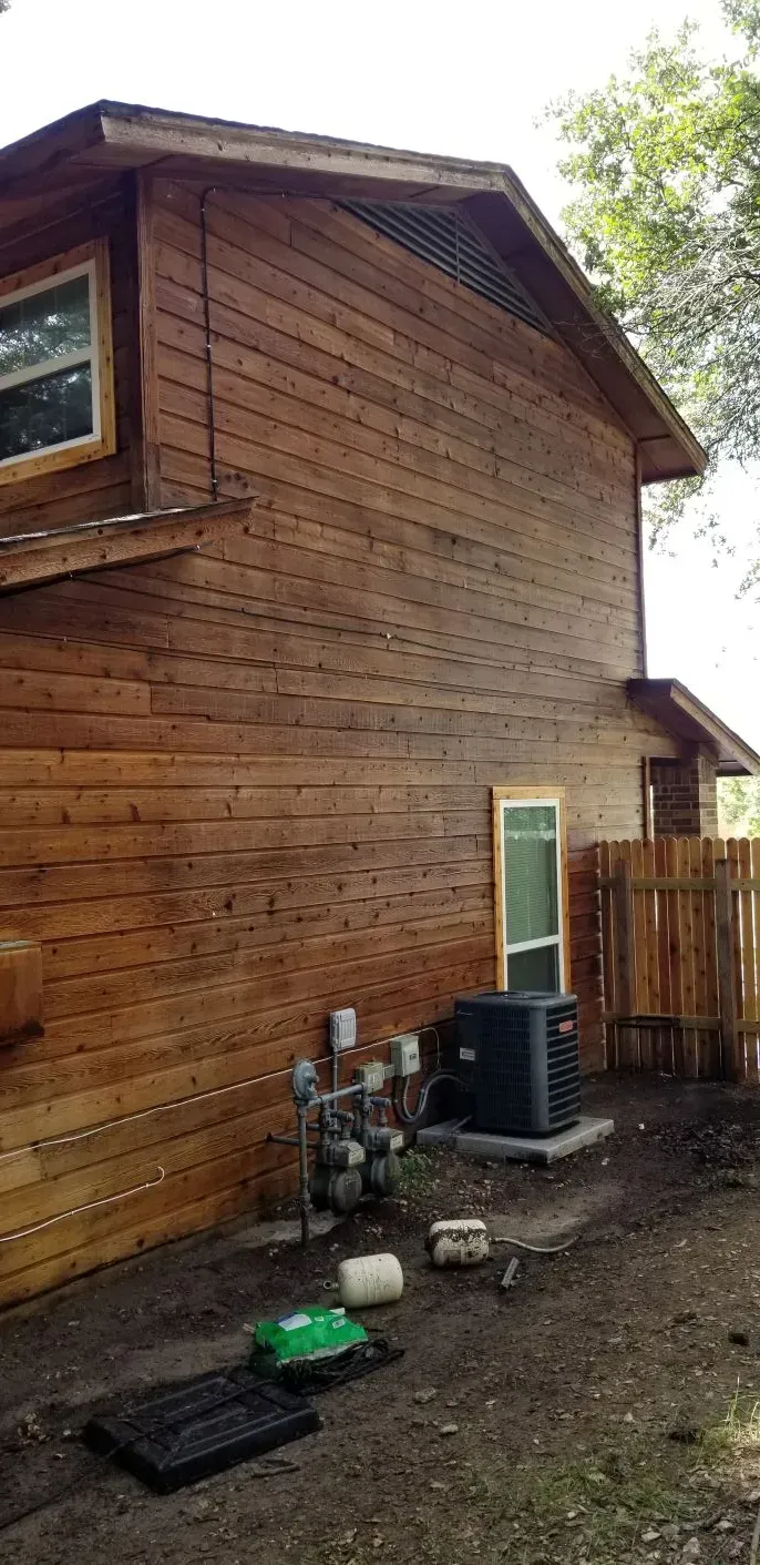 Wooden-sided two-story house with an air conditioning unit and fencing in a grassy backyard.
